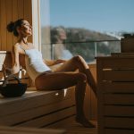Eine Frau in einem weißen Badeanzug entspannt sich auf einer Holzbank in einer sonnenbeschienenen Sauna. Sie hat die Augen geschlossen und genießt die Wärme, während sie durch ein großes Fenster den Blick nach draußen schweifen lässt.