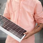 Person in einem pfirsichfarbenen Hemd, die mit beiden Händen ein kleines rechteckiges Solarpanel hält; nur der Oberkörper ist sichtbar, ein Unterarm ist tätowiert.
