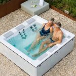 Ein lächelndes älteres Ehepaar entspannt sich in einem modernen Whirlpool im Freien auf einer Kiesfläche, umgeben von Grünflächen. Handtücher, Getränke und Sonnenbrillen liegen in der Nähe auf einem Holzsockel.