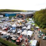 Luftaufnahme einer Freiluftveranstaltung mit vielen Zelten, Ständen und Fahrzeugen. Die Menschen flanieren zwischen den Auslagen auf einem großen Parkplatz in der Nähe von Industriegebäuden und Grünanlagen, im Hintergrund sind ein MediaMarkt und "Pressemitteilung"-Banner zu sehen.