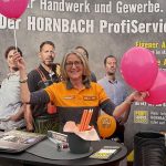 Eine lächelnde Frau in einer Hornbach-Uniform steht an einem Stand und hält rosa Luftballons in der Hand. Hinter ihr hängt ein Banner mit der Aufschrift HORNBACH ProfiService und Bildern von Menschen in Arbeitskleidung. Auf dem Tisch liegen Flyer und Bleistifte.