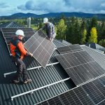 Zwei Arbeiter mit Helmen installieren Solarpaneele auf einem Metalldach in einer ländlichen Gegend mit Bäumen und Bergen im Hintergrund unter einem bewölkten Himmel.