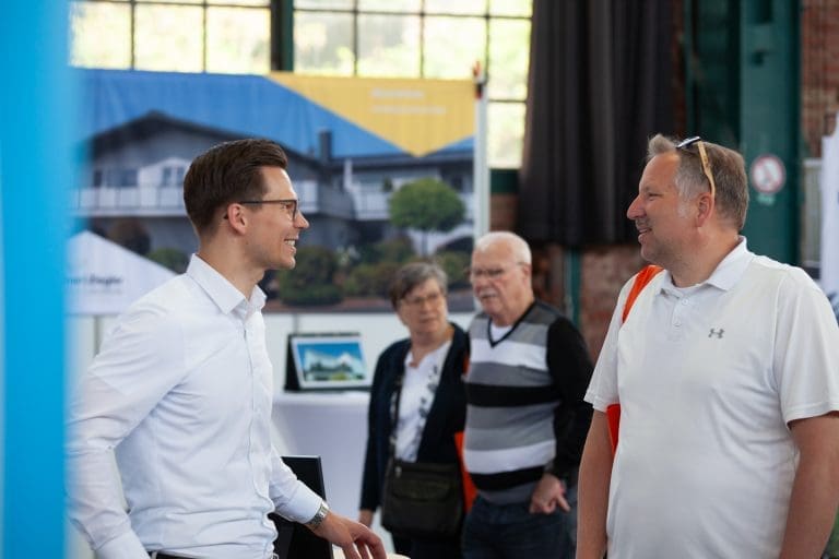 Zwei Männer stehen und unterhalten sich bei einer Indoor-Veranstaltung, beide lächeln. Drei Personen im Hintergrund beobachten Auslagen. Hinter ihnen sind leuchtende Transparente und ein gerahmtes Foto eines Hauses zu sehen. Alle sind leger gekleidet.