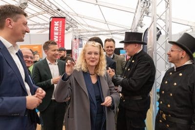 Eine Frau in einem grauen Mantel lächelt und hält einen kleinen Gegenstand in der Hand, während sie zwischen mehreren Männern, einige in Anzügen, andere in schwarzen Uniformen mit Zylinder, in einem Zelt auf einer Veranstaltung oder Ausstellung steht.