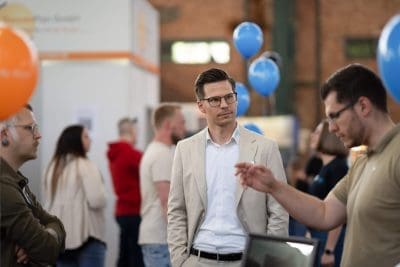 Ein Mann in einem beigen Anzug steht inmitten von Menschen bei einer Indoor-Veranstaltung mit blauen und orangefarbenen Luftballons im Hintergrund. Andere Teilnehmer unterhalten sich und gehen umher, und die Umgebung wirkt locker und gesellig.