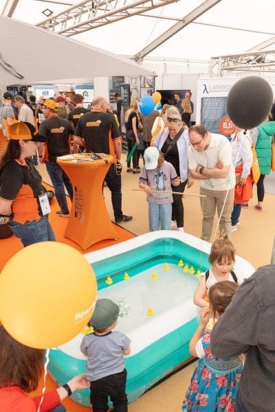 Menschen versammeln sich bei einer Indoor-Veranstaltung um einen kleinen aufblasbaren Pool mit schwimmenden gelben Spielzeugenten. Kinder spielen in der Nähe des Beckens, während Erwachsene zusehen und sich unterhalten. Einige Leute halten orangefarbene und schwarze Luftballons; im Hintergrund sind Stände und Auslagen zu sehen.