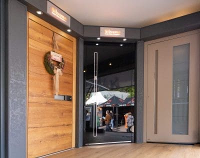 Ein Ausstellungsraum mit zwei modernen Türen; die linke Tür ist aus Holz mit einem Kranz, die rechte Tür ist beige, und durch die Glastür in der Mitte kann man die Leute draußen sehen. Auf den Schildern darüber steht Silvanus.