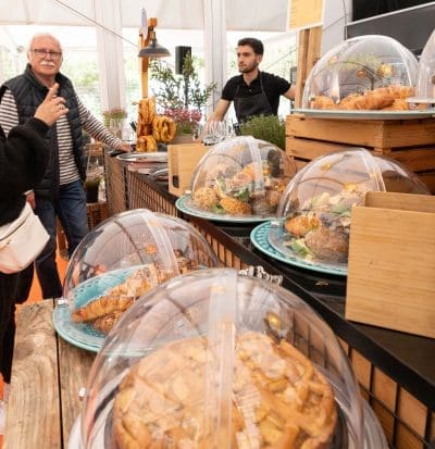 Ein Bäckereimarktstand bietet unter durchsichtigen Kuppeln verschiedene Backwaren an. Ein Verkäufer steht hinter der Theke, während ein Mann und eine Frau davor die Produkte auszuwählen scheinen. Die Einrichtung ist rustikal mit Holzkisten und Grünzeug.